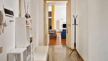 Un couloir avec un banc blanc, un porte-manteau et une porte donnant sur un salon équipé d’un canapé et d’un téléviseur.