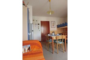 Quarto com uma cama, uma mesa de jantar com cadeiras, um refrigerador, uma porta de madeira e uma prateleira fixada na parede.