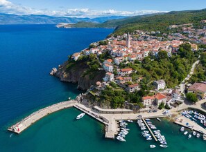 An aerial view of a coastal town with a marina, buildings with red-tiled roofs, and a prominent church tower.