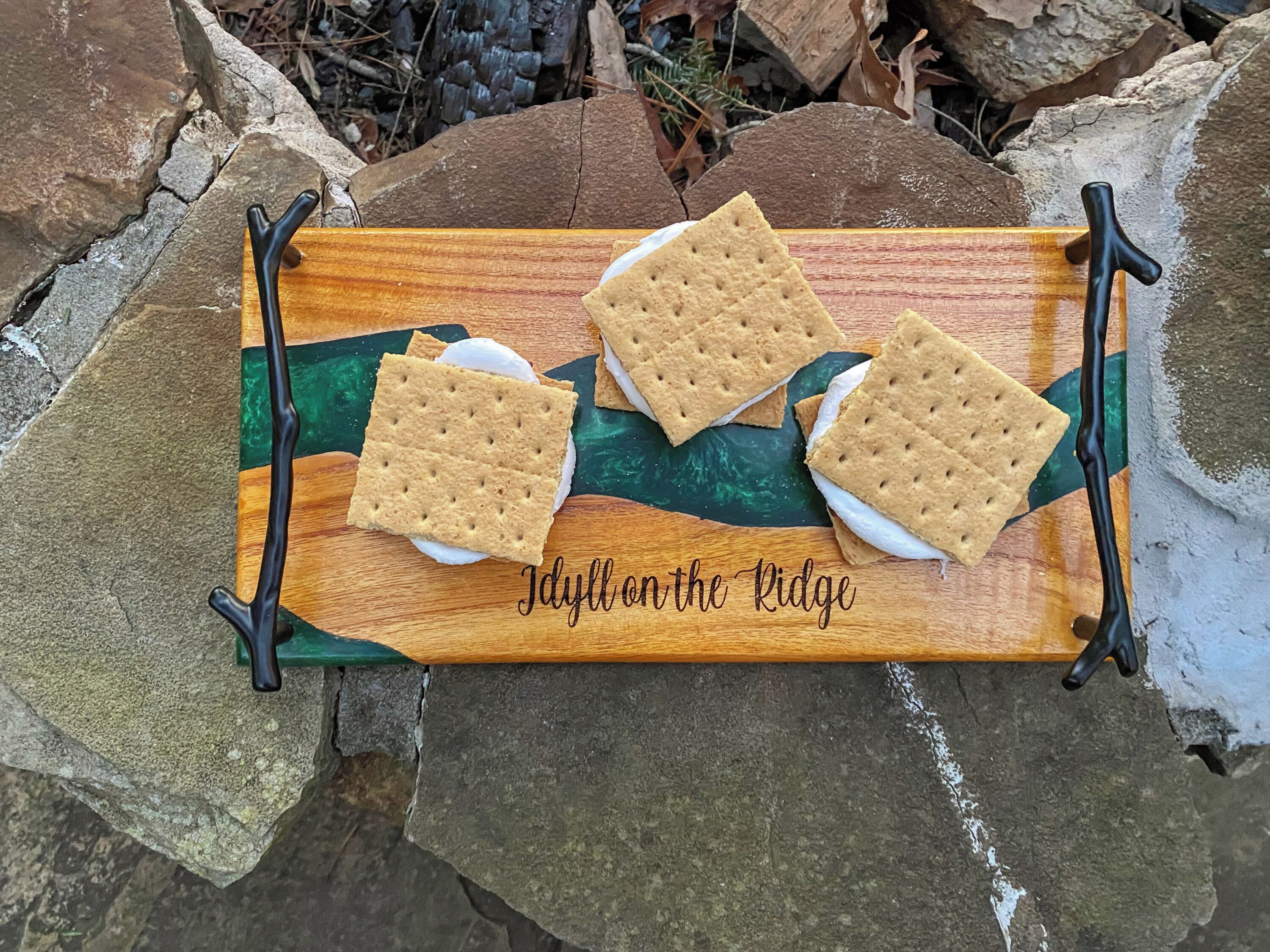 Un plateau en bois avec trois « s’mores » posés sur une surface en pierre.