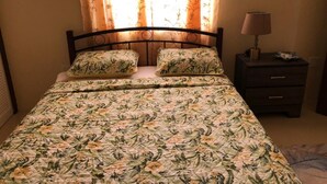 A bedroom with a floral bedspread, two matching pillows, a nightstand with a lamp, and a window with curtains. - Jomp Turners' Guest Apartments (Bolands)