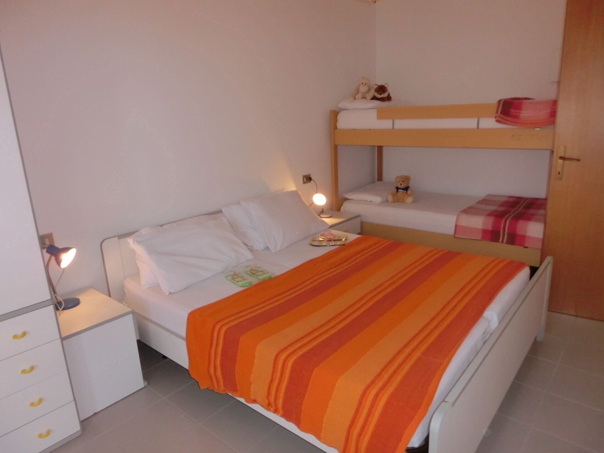 Un dormitorio con una cama litera, una mesita de noche con lámpara, un aparador con cajones y una manta sobre la cama.