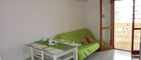 A small room with a green sofa, a white table with chairs, and a sliding glass door leading to an outdoor area.