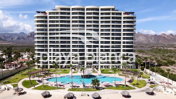 A multi-story hotel with a pool area, palm trees, and mountains in the background.