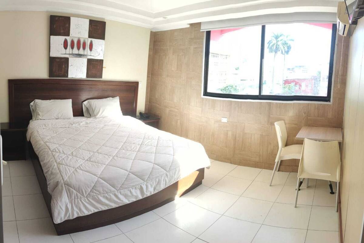Une chambre avec un grand lit, une tête de lit en bois, une petite table avec une chaise et une fenêtre donnant sur des palmiers.