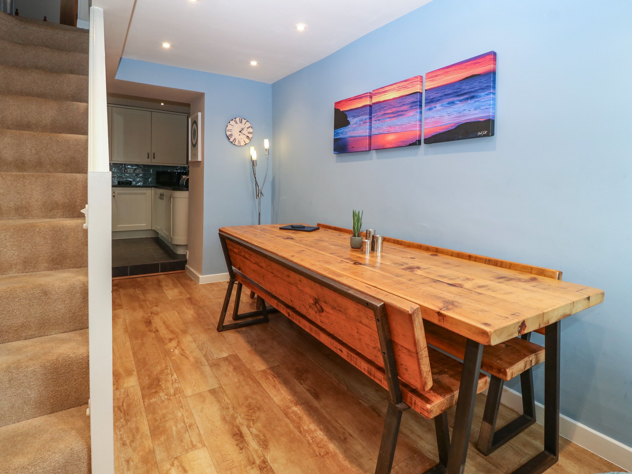 Un comedor con una mesa de madera y bancos, un reloj y un cuadro en la pared.