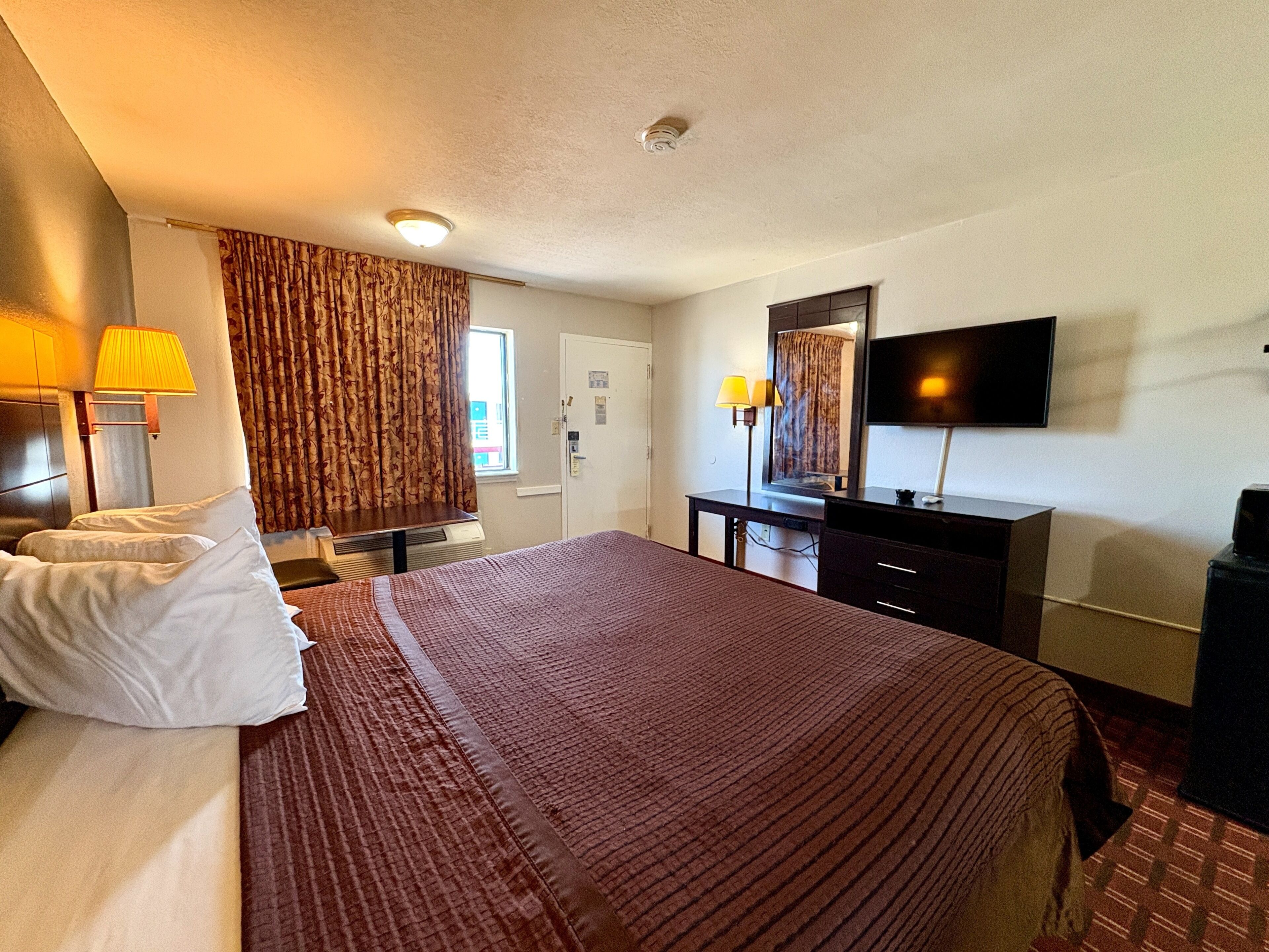 Habitación de hotel con cama, escritorio, televisor y ventana con cortinas.