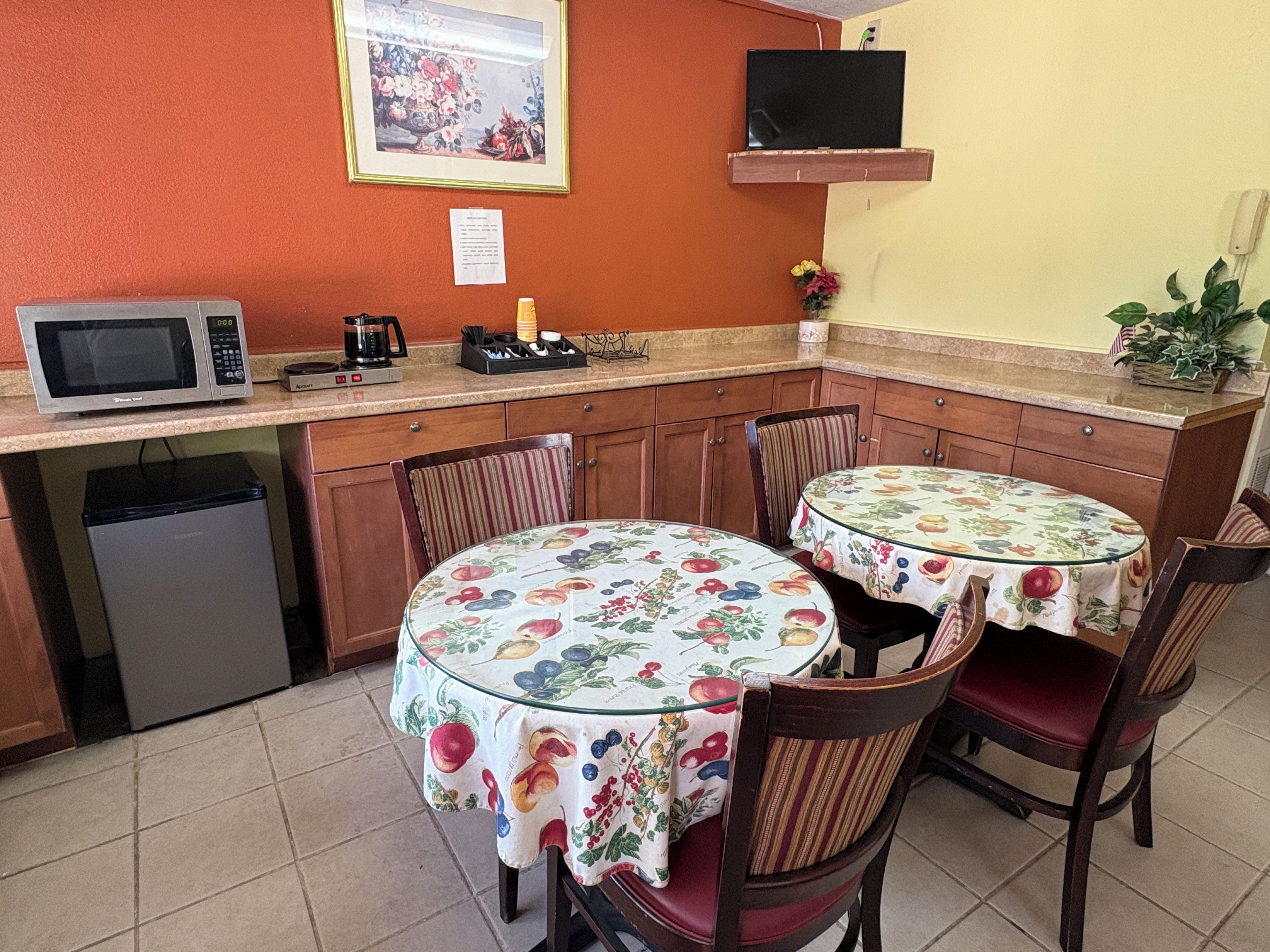 A compact kitchen with a microwave, coffee maker, and a small refrigerator. There are two dining tables with chairs, one of which has a floral tablecloth. A television is mounted on the wall above the kitchen counter.