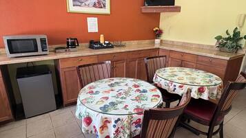 A compact kitchen with a microwave, coffee maker, and a small refrigerator. There are two dining tables with chairs, one of which has a floral tablecloth. A television is mounted on the wall above the kitchen counter.