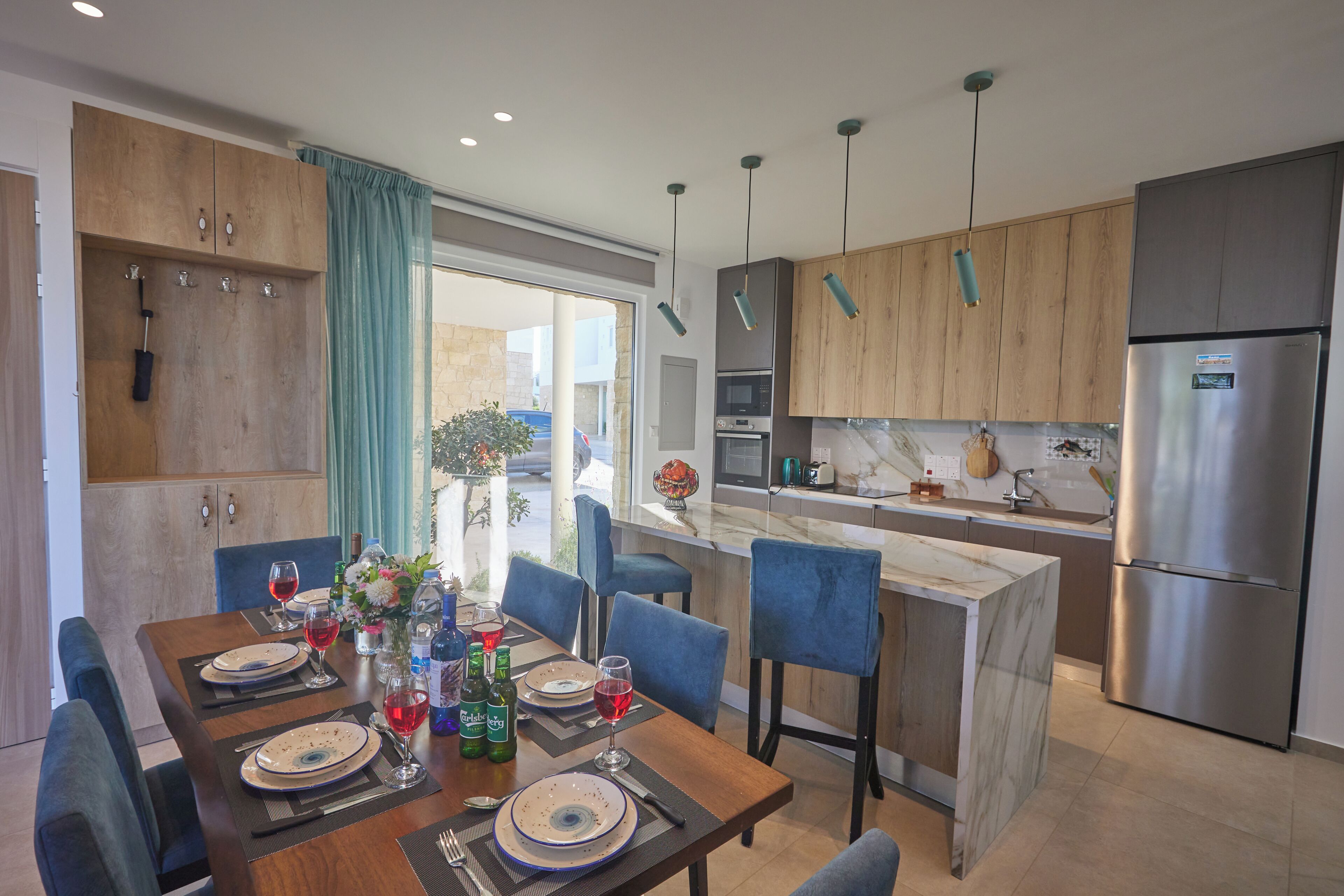 A modern kitchen with wooden cabinets, a stainless steel refrigerator, and a dining area with blue chairs and a marble table.