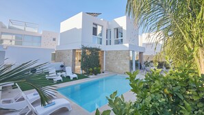 A modern poolside area with white lounge chairs, a clear blue pool, and a stone building with large windows. - Protaras Vie Bleu Villa Vb8 (Paralimni)