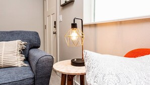 Una habitación acogedora con un sofá gris, una pequeña mesa redonda con una lámpara y una cama con almohadas estampadas.