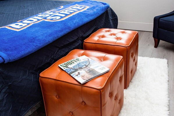 Una camera da letto con un letto ricoperto da una coperta blu, due pouf in pelle marrone e una rivista su uno dei pouf.