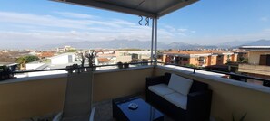 Een balkon met een bank en tafel, met uitzicht op een woongebied met huizen en bergen in de verte.
