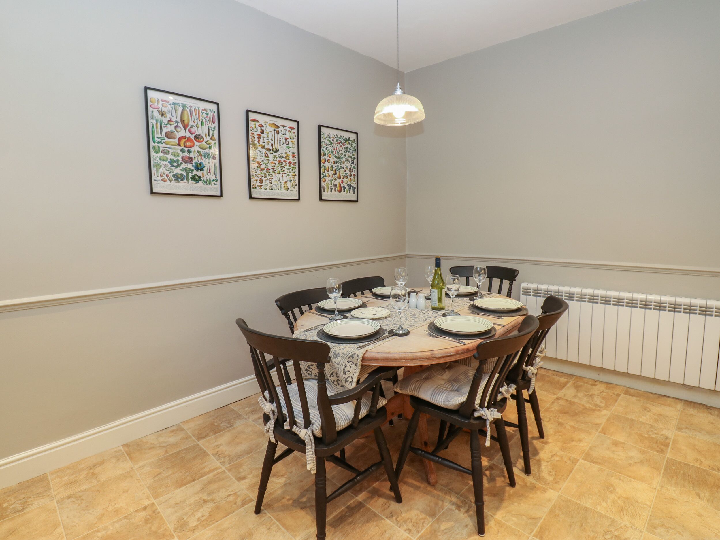 Een eetruimte met een ronde tafel voor zes personen, omringd door houten stoelen met beklede zitplaatsen. Boven de tafel hangen drie ingelijste botanische afbeeldingen.