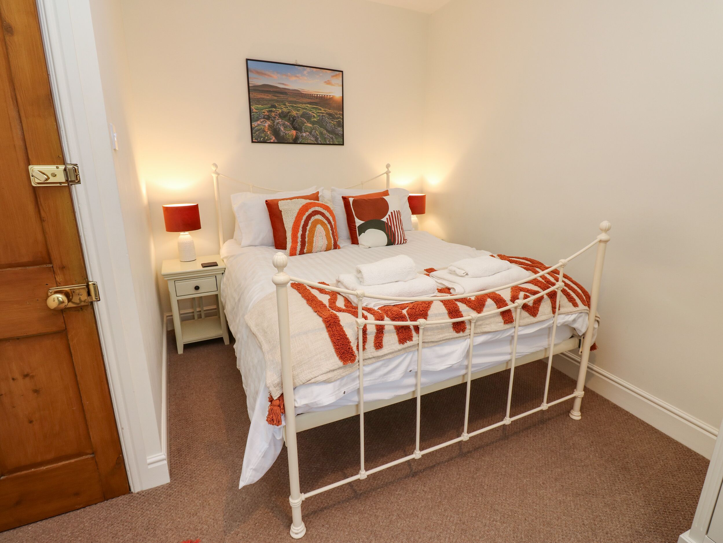 A neatly made bed with white bedding and orange accents, a bedside table with a lamp, and a painting on the wall.