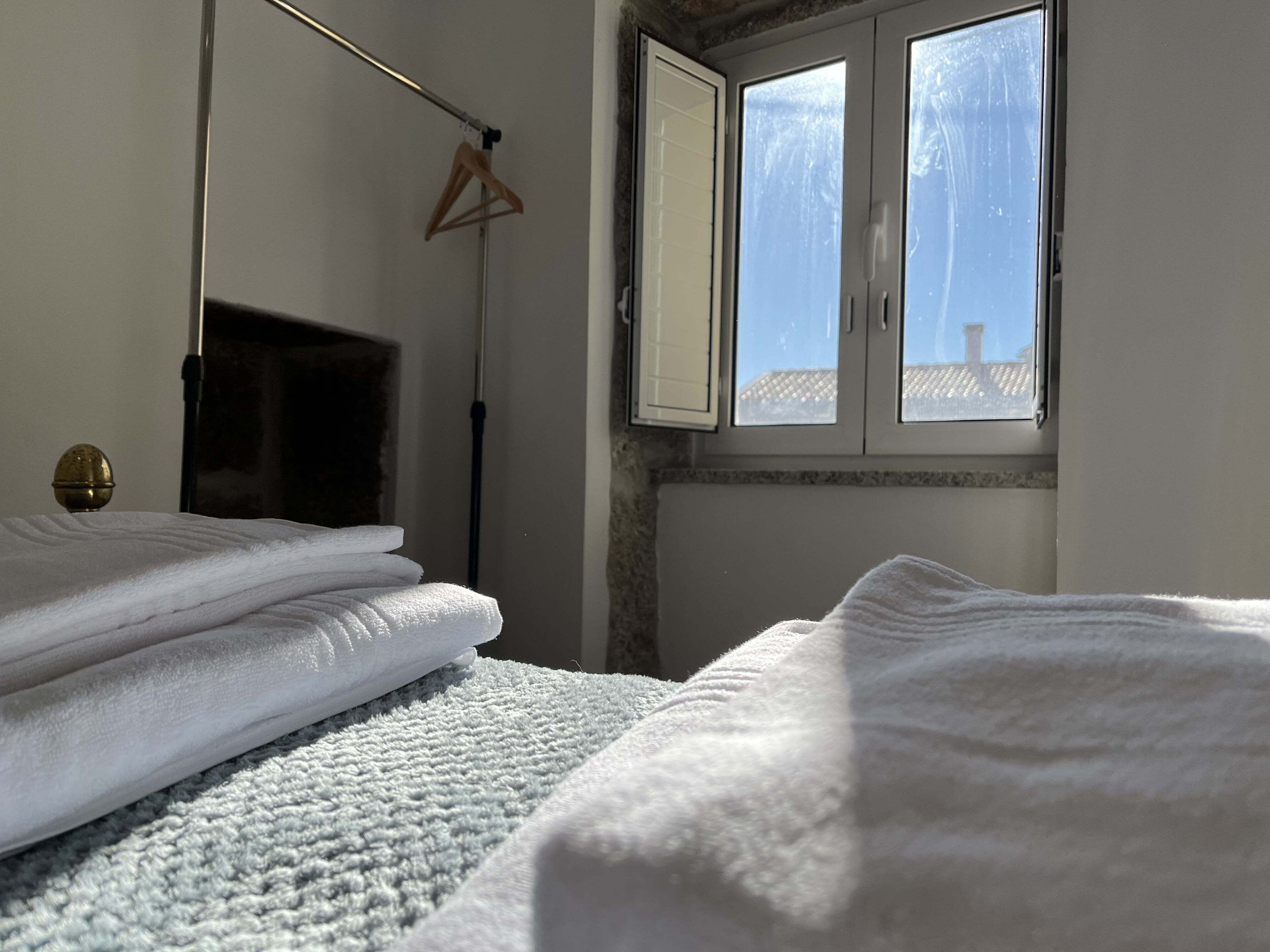 Ein ordentlich bezogenes Bett mit weißen Leinen, ein hölzerner Kleiderbügel an einer Stange und ein Fenster mit Blick auf den Himmel und ein Gebäude.