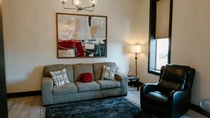 A living room with a grey sofa, a black leather chair, a patterned rug, a floor lamp, and a large abstract painting on the wall.
