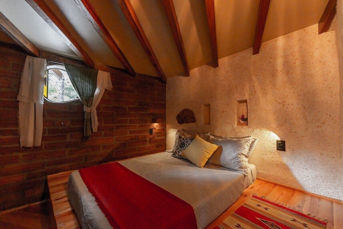 Um quarto aconchegante com parede de tijolos, vigas de madeira, cama com roupa de cama branca e um cobertor vermelho, além de uma pequena janela com cortinas.