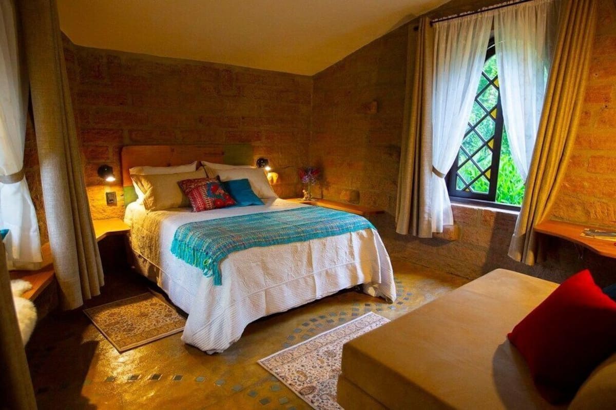 Um quarto aconchegante com uma cama grande, cabeceira de madeira, mesa de cabeceira com abajur e uma janela com cortinas.