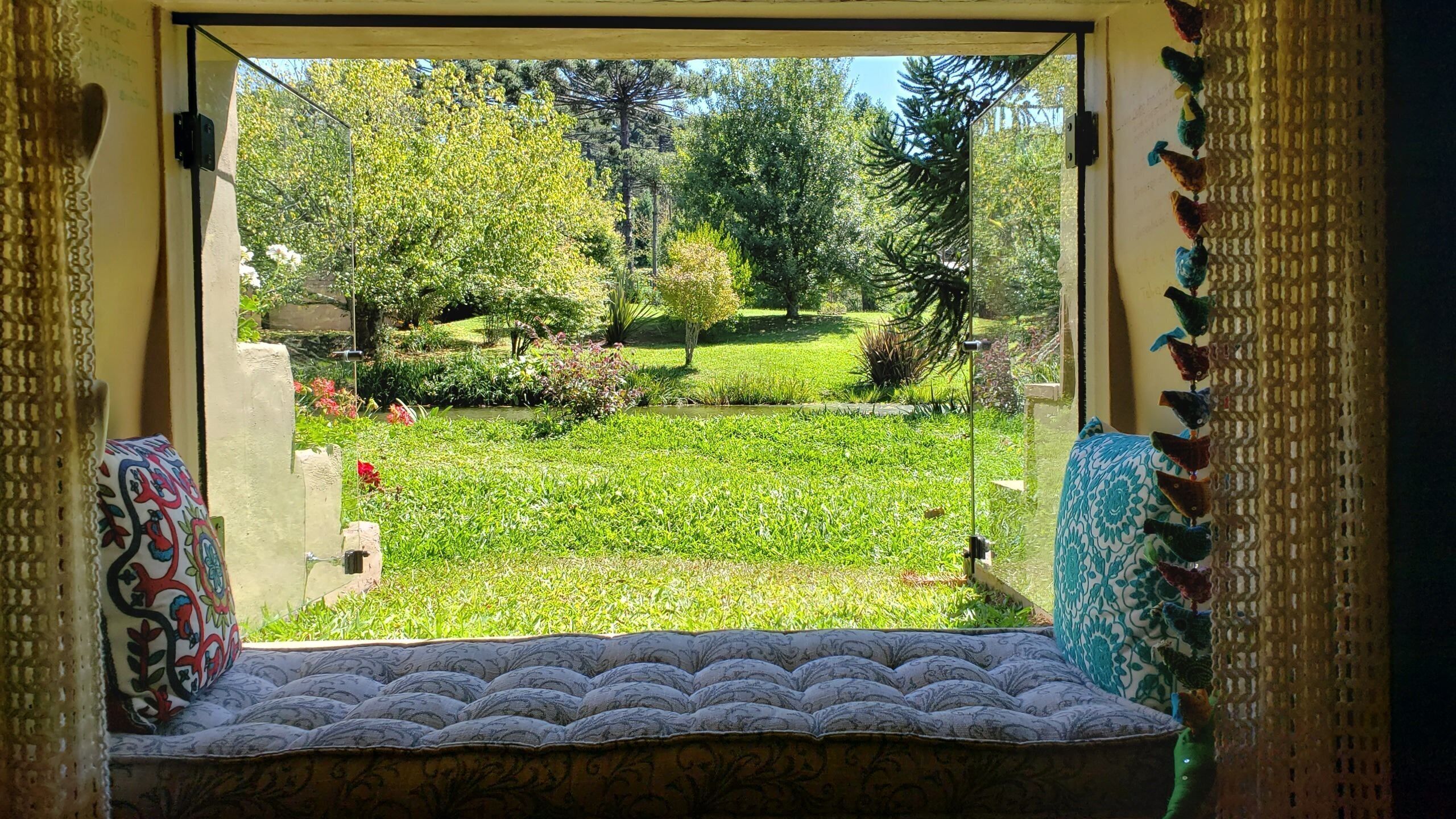 Une fenêtre donnant sur un jardin, un banc et un coussin à motifs.