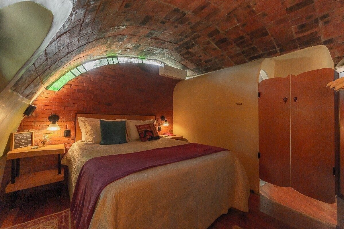 Quarto aconchegante com teto de tijolos, uma cama com almofadas, uma mesa de cabeceira com abajur e um guarda-roupa.