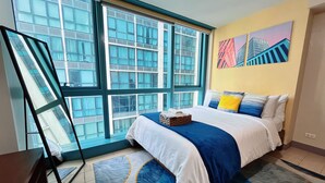 A modern hotel room with a large window, a bed with blue and white bedding, a blue and white rug, and a painting on the wall. - LARGE BGC 2BR, Steps to Mitsukoshi & Uptown Malls, 300Mbps Fiber 25R (Taguig)