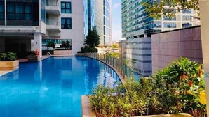A swimming pool surrounded by modern buildings with balconies, a clear sky, and some greenery. - LARGE BGC 2BR, Steps to Mitsukoshi & Uptown Malls, 300Mbps Fiber 25R (Taguig)