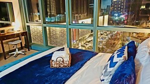 Ein Hotelzimmer mit einem großen Bett, einem Schreibtisch und einem Sessel. Ein Fenster bietet Blick auf eine nächtliche Stadtansicht.