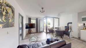 Ein modernes Wohnzimmer mit Flachbildfernseher, Glastisch und Sofa. Es gibt einen Essbereich mit Tisch und Stühlen sowie eine Küchenzeile mit Kühlschrank und Kaffeemaschine.