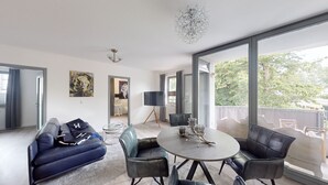 Ein modernes Wohnzimmer mit einer Couch, einem runden Tisch und Stühlen. Ein großes Fenster bietet Blick auf Bäume und einen Balkon.
