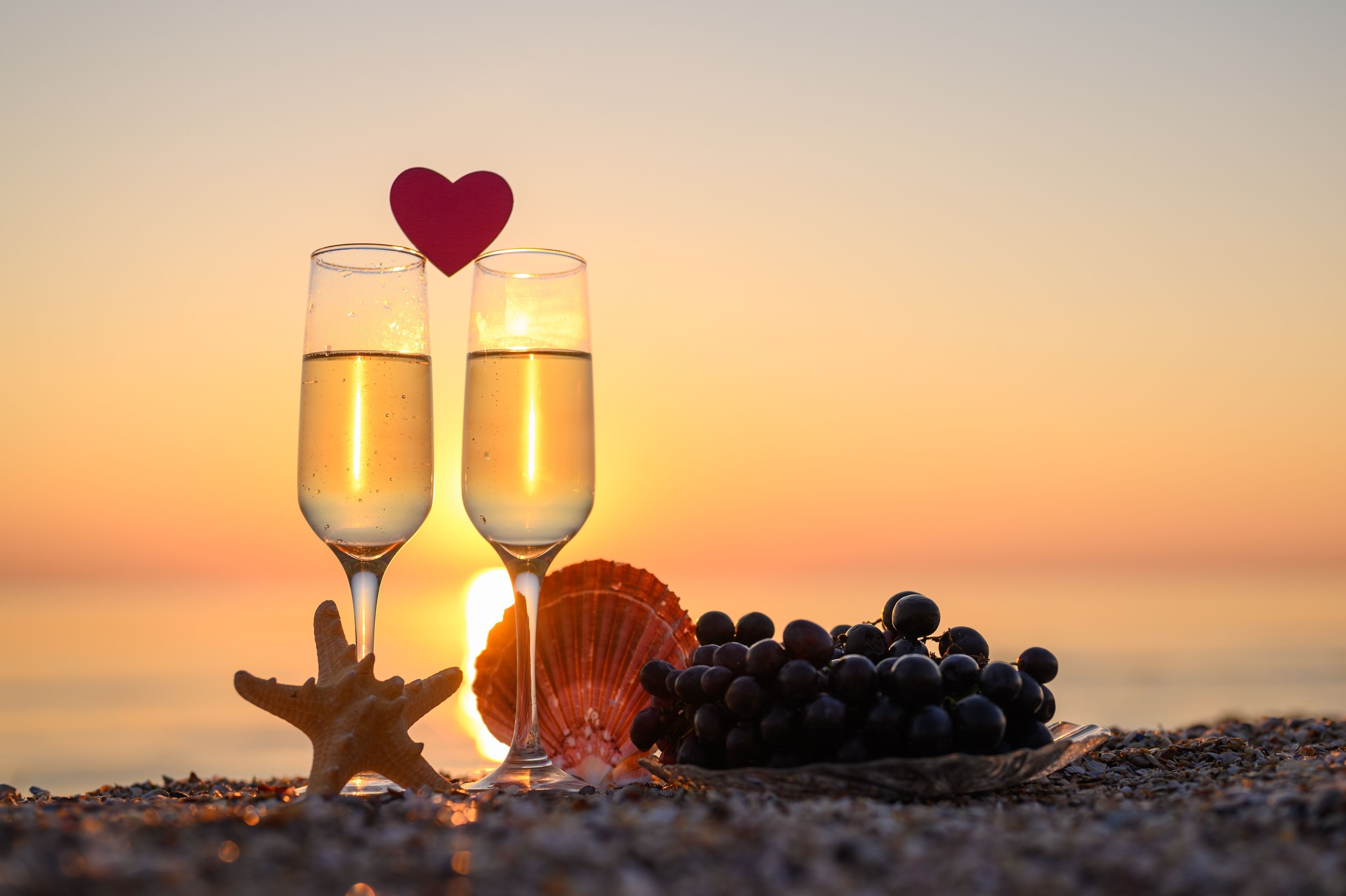 Two champagne flutes with a heart-shaped decoration, a starfish, a scallop shell, and a bunch of grapes on a beach at sunset.