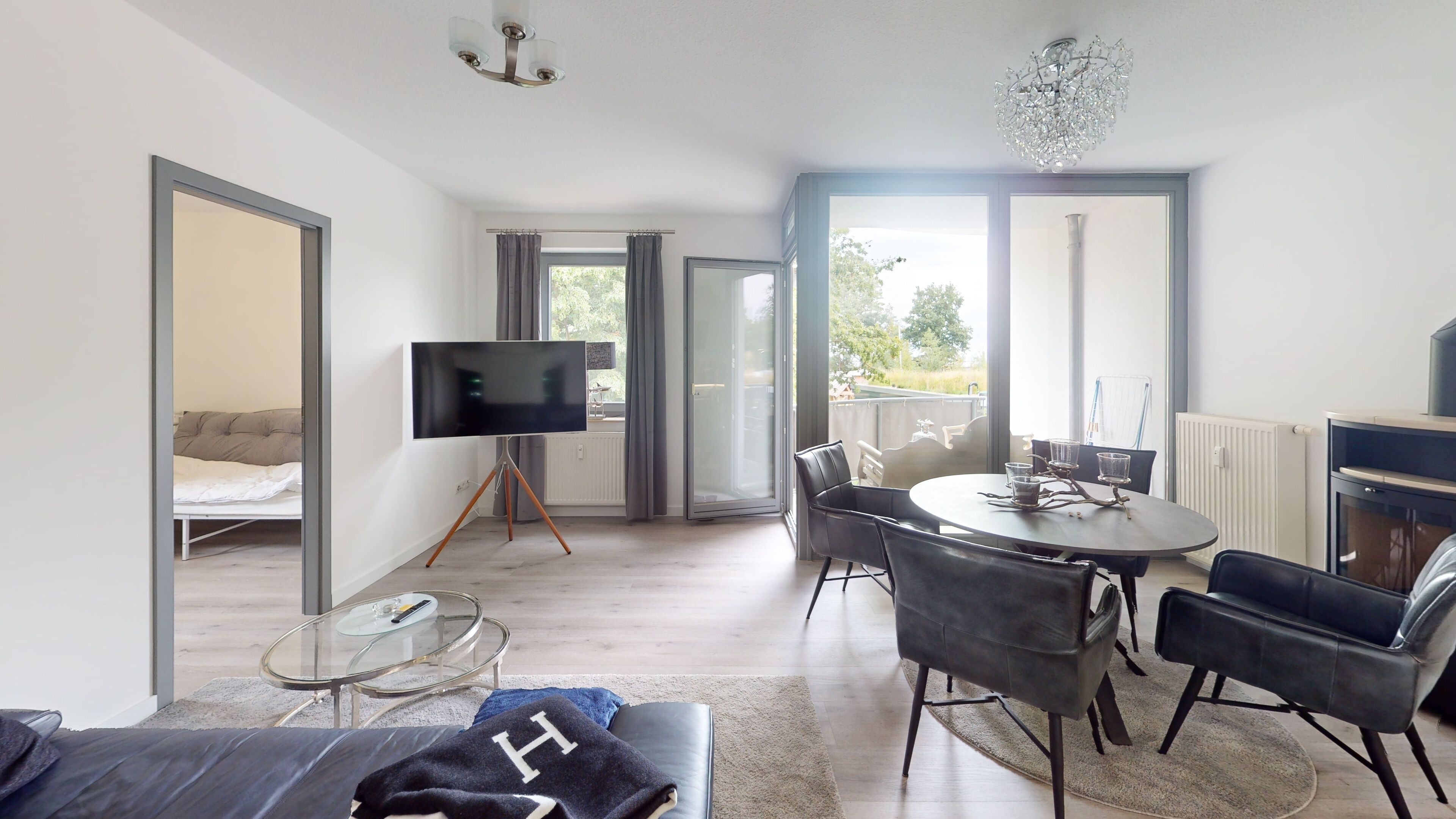 A modern living room with a sofa, a glass coffee table, a dining table with chairs, a flat-screen TV, and a chandelier.