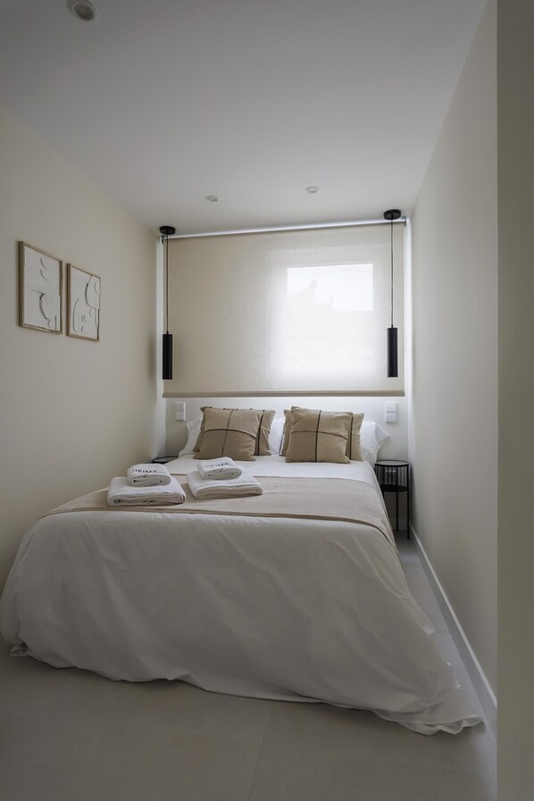 A neatly made bed with a white duvet and two pillows, a bedside table with a lamp, and a window with blinds.