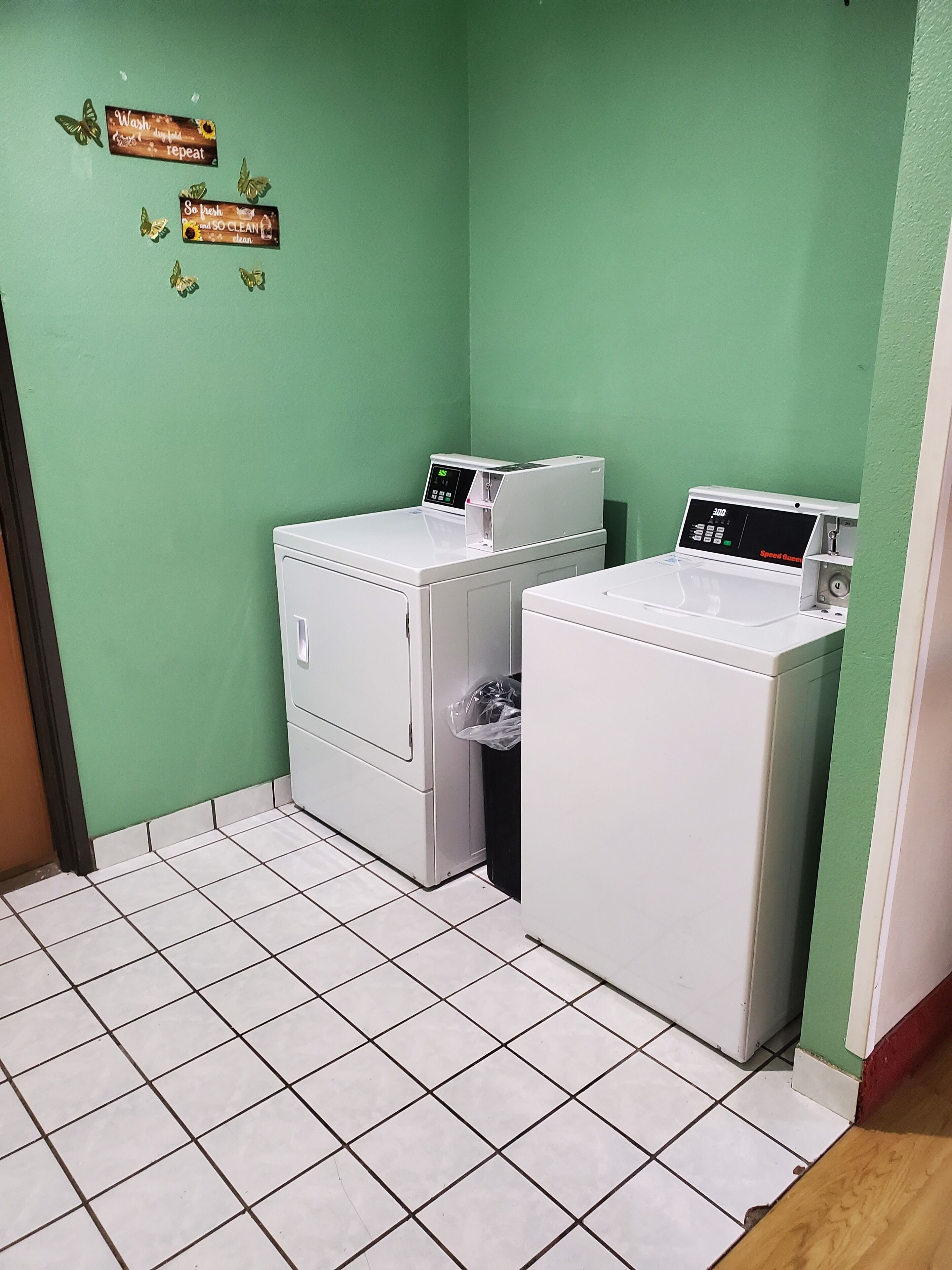 A laundry room with two white front-loading washing machines, a trash bin, and a green wall with motivational signs.