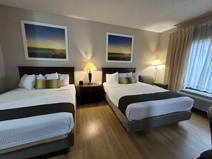 A hotel room with two beds, a nightstand with a lamp, a window with curtains, and two landscape paintings on the wall. - McPherson Inn (McPherson)