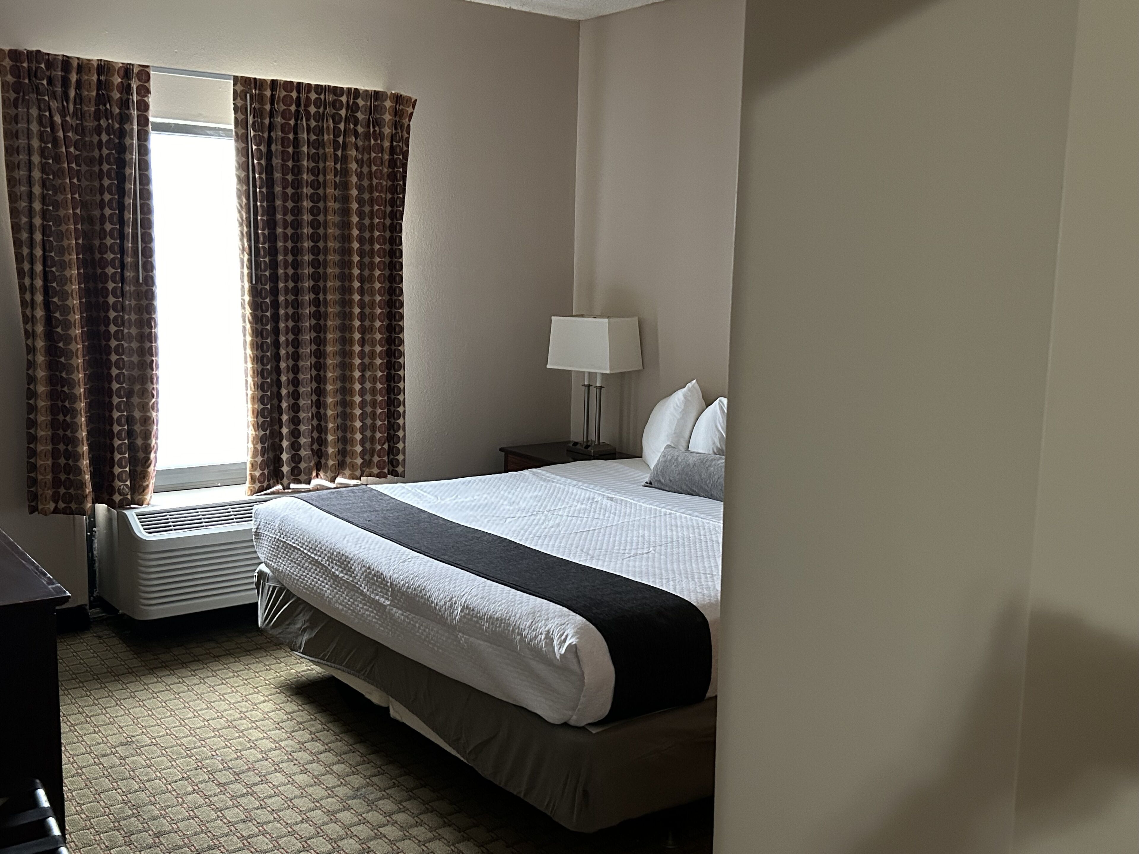 Una habitación de hotel con una cama, una mesita de noche, una lámpara y una ventana con cortinas.