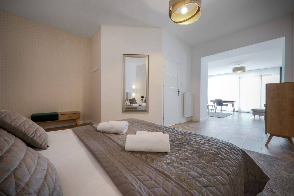 A modern hotel room with a large bed, a mirror, and a view of the dining area.