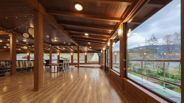 A spacious indoor area with wooden floors and large windows offering a view of a lake and trees. - Hotel Łaziska (Czchow)