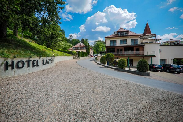 La entrada de un hotel con un camino de grava, un cartel que dice "HOTEL LAZARUS" y un edificio con techo rojo y balcón.