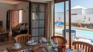 Un comedor con una mesa preparada para cuatro personas, vista a una piscina y puerta que da a una sala de estar.