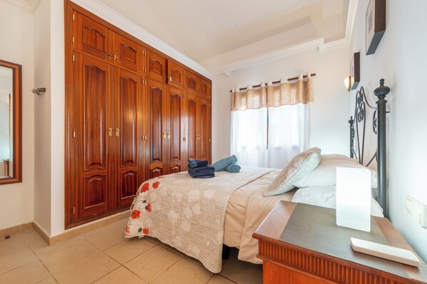 Une chambre à coucher avec une grande armoire, un lit recouvert d’une couvre-lit à motifs floraux et une table de chevet avec une lampe.