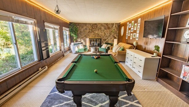 Une table de billard dans une pièce dotée de grandes fenêtres, d’un mur en pierre et d’un téléviseur fixé au mur.