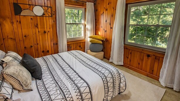 Ein Zimmer mit holzverkleideten Wänden, einem Bett mit gemusterten Betttextilien, zwei Fenstern mit Vorhängen und einem kleinen Nachttisch mit Lampe.