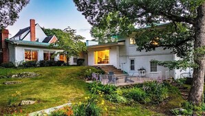 Une maison à deux étages avec un porche à l’avant, un escalier apparent et un jardin bien entretenu où poussent différentes plantes et fleurs.