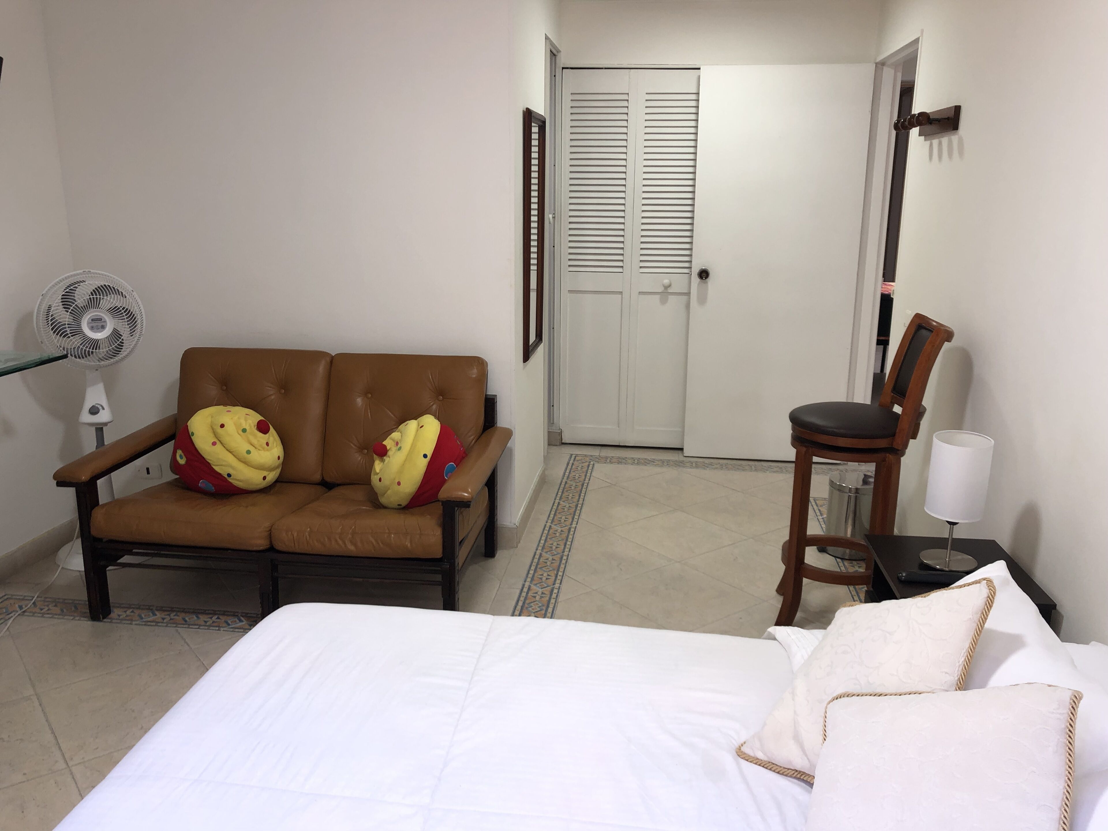 A hotel room with a bed, a brown leather sofa with two large cupcake-shaped pillows, a wooden chair, a small table with a lamp, and a white door in the background.