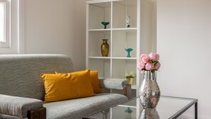 A living room with a grey sofa, a yellow pillow, a glass coffee table, and a white shelving unit with decorative items.