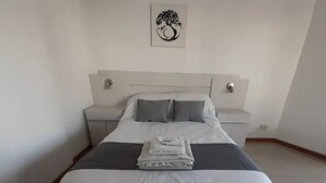 Un letto ben rifatto con copripiumino bianco e grigio, un asciugamano piegato e una testiera con un'opera d'arte a parete.