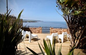 Una terraza de madera con sillas de plástico blancas, un muro de ladrillo y vista al mar.