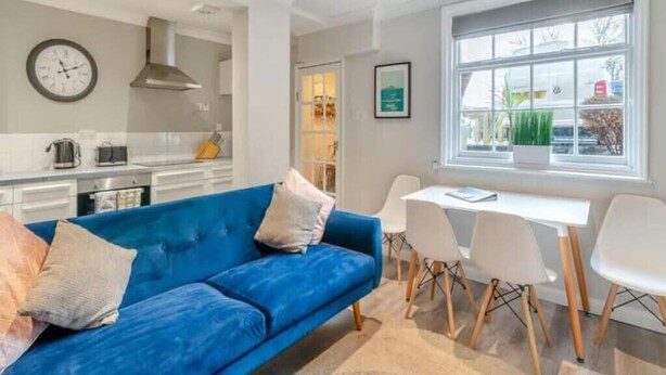 A modern living room with a blue sofa, white chairs, a dining table, and a large window with a view of the outside. - Molokai Snug | Best location | North South bay | Courtyard garden | Parking (North Yorkshire)